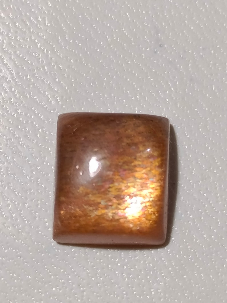 Op de afbeelding: Een vierkante, iriserende sunstone cabochon met een roodbruine kleur en een glinsterende, glimmende afwerking.