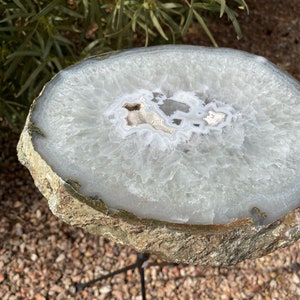 May include: A large, white agate slice with a natural, rough edge, mounted on a black metal tripod stand. The agate has a white and gray pattern with a clear center.