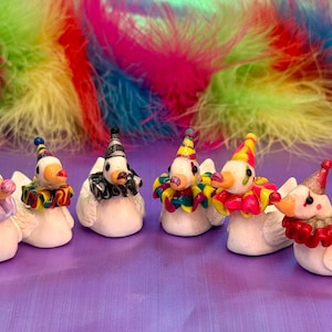 May include: Six white duck figurines, each adorned with a clown hat and ruffled collar in various colors. The ducks have painted eyes and beaks, set against a backdrop of colorful feathers. A whimsical and decorative item.