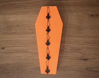 May include: A black coffin-shaped ornament with a row of five black cross charms hanging from it. The charms are attached to the ornament with silver jump rings.