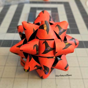 May include: A red and black starburst bow with a witch hat pattern. The bow is made of paper and has a 3D effect. The text "StarboardBows" is visible in the bottom right corner of the image.