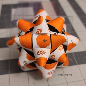 May include: A white and orange paper bow with a pumpkin and leaf design. The bow is folded into a star shape.