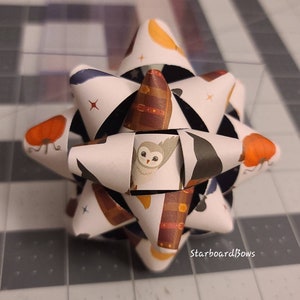 May include: A decorative paper bow with a repeating pattern of pumpkins, owls, and books. The bow is white with black accents and has the text "StarboardBows" printed on it. The bow is a festive decoration.