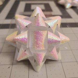 May include: A white iridescent bow with a starburst shape. The bow is made of paper and has a textured surface.
