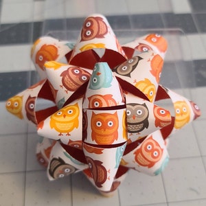 May include: A white paper bow with a red center and a pattern of owls in orange, brown, blue, and white.