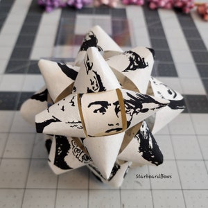 May include: A white paper starburst ornament with a black and white print of a woman's face. The ornament is folded into a 3D shape and has a starburst design. The text "StarboardBows" is printed on the bottom of the ornament.