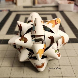 May include: A white paper starburst bow with a dog print design. The bow features various breeds of dogs, including a husky, a dachshund, and a golden retriever. The text "Starboard Bows" is printed on the bottom of the bow.