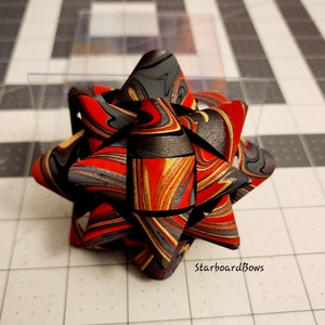 May include: A red, black, and gold marbled starburst bow. The bow is made of polymer clay and has a swirl pattern.