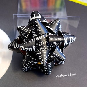 May include: A decorative bow made of black paper with white text. The bow is in a starburst shape, with the text wrapping around the curves of the bow. The bow is sitting on a gray surface.