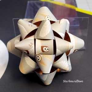 May include: A brown and white paper gift bow with a repeating pattern of owls. The bow is shaped like a star and has a textured surface.