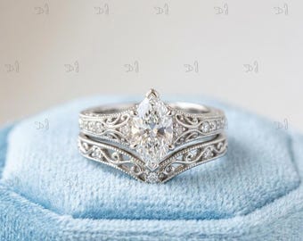 May include: A silver-toned engagement ring set featuring a marquise-cut center stone. The band and the matching contoured wedding band both display intricate filigree metalwork and small round accent stones along the shank of the rings.