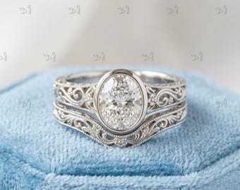 May include: A silver-toned ring set featuring an oval-cut center stone held in a bezel setting. The band and the matching contoured wedding band are decorated with intricate scrollwork and milgrain detailing along the metal surfaces.