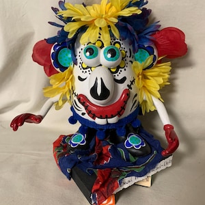 May include: A Day of the Dead-themed decorative figurine. The figure has a white face with black details, large blue eyes, and a red mouth. It is adorned with yellow and blue floral accents and wears a blue and red patterned outfit.