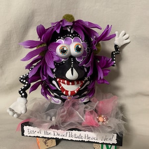 May include: A Day of the Dead Potato Head figure, painted black with white accents, purple flower hair, and a red and white smile. The figure has white hands with black polka dot arms and is on a black base with the text "Day of the Dead Potato Head-Ned".