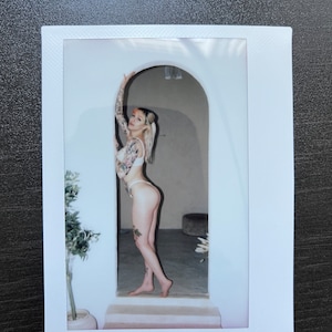 May include: Polaroid-style photograph of a person in lingerie, posed in an arched doorway. The person has tattoos on their arms and legs. The image is framed by a white border, with a dark surface in the background.
