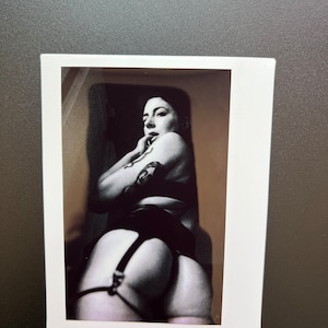 May include: A black and white photograph of a person wearing a black suspender belt and stockings. The person is looking over their shoulder and has a tattoo on their arm.