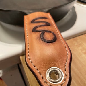 May include: A brown leather handle with a stitched edge and a metal rivet. The handle has the word "DC" embossed on it.