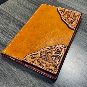 May include: A brown leather journal with intricate floral tooling. The design features a large flower in the center and smaller flowers and leaves around it.