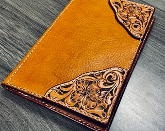 Puede incluir: Un diario de cuero marrón con intrincados detalles florales. El diseño presenta una flor grande en el centro y flores y hojas más pequeñas alrededor.
