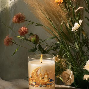 May include: A lit candle in a clear glass jar, featuring a gold crescent moon and lantern design. The candle is surrounded by flowers and foliage, creating a warm and inviting ambiance. The candle's flame is bright, and the overall aesthetic is elegant.