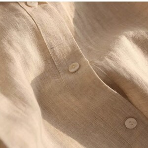 Pu&ograve; includere: Primo piano di una camicia di lino beige con chiusura a bottoni. Il tessuto ha una texture sottile e un aspetto naturale e rilassato.