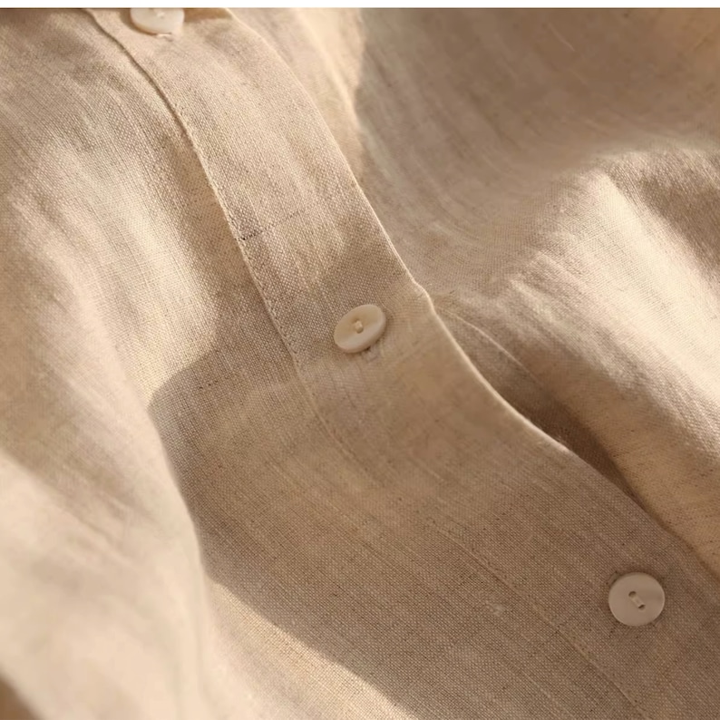 Pu&ograve; includere: Primo piano di una camicia di lino beige con chiusura a bottoni. Il tessuto ha una texture sottile e un aspetto naturale e rilassato.