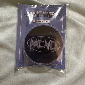 May include: A black Smart Tok phone grip with the word "MCND" in white, stylized lettering. The grip is packaged on a blue card with the words "ADD VENTURE" and "SMART TOK" printed on it. The packaging is clear plastic.