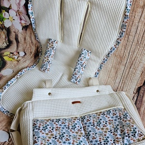 Può includere: Un set color crema per neonati composto da inserto per passeggino, coprimaniglie e borsa per pannolini. Il set presenta una trama a nido d'ape e dettagli floreali blu, marroni e verdi. La borsa per pannolini ha due tasche frontali.