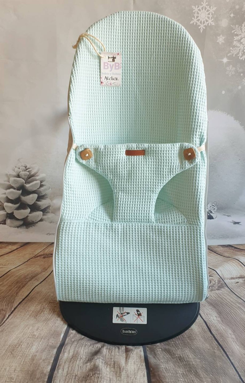 May include: A light green waffle weave fabric baby chair cover with a brown leather tag that says "ByB Atelier". The cover is on a black base with the BumBo logo.