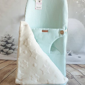 May include: A light blue waffle weave baby nest with a white plush blanket with star and flower patterns. The nest has a brown leather strap and a wooden button. The tag on the nest says "ByB Atelier".
