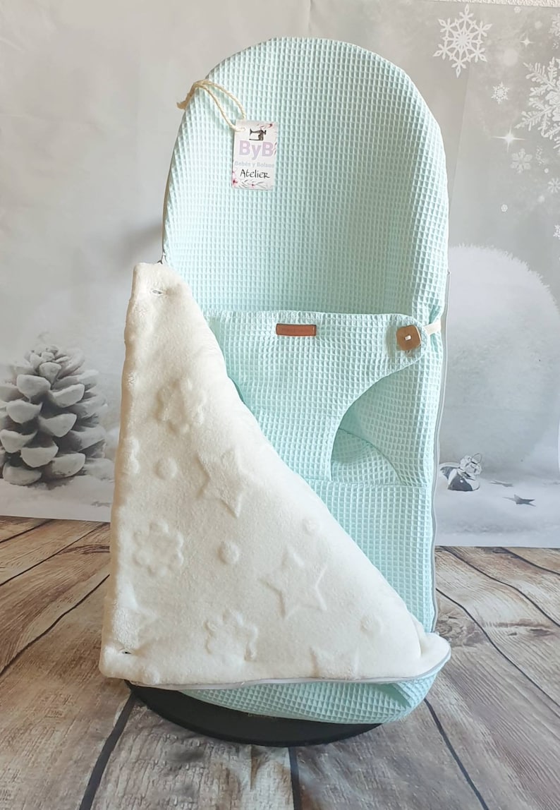 May include: A light blue waffle weave baby nest with a white plush blanket with star and flower patterns. The nest has a brown leather strap and a wooden button. The tag on the nest says "ByB Atelier".