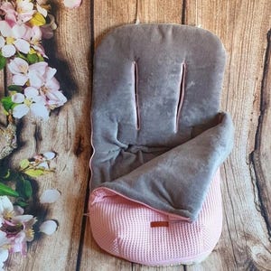Peut inclure: Un nid d'ange gris et rose pour poussette. La partie supérieure est en matériau gris doux avec une bordure rose et deux fentes verticales. Le bas est en tissu rose texturé. Une petite étiquette marron est cousue sur le devant.