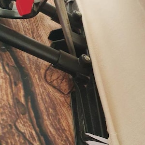 May include: Close-up of a black metal frame with a white fabric cover. The frame has a red lever and a black metal bar. The fabric cover is attached to the frame with white elastic bands.