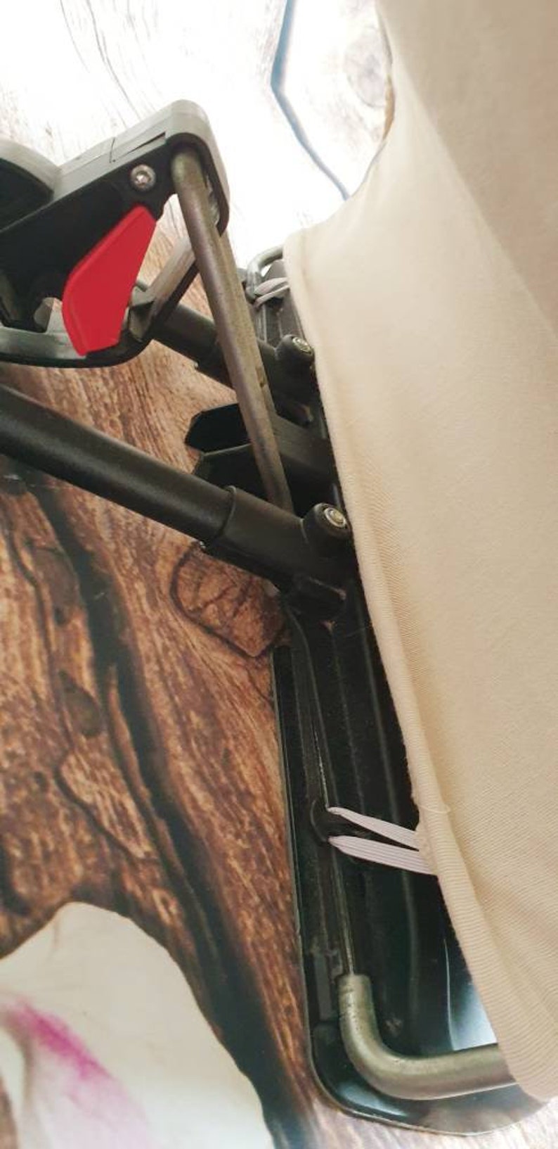 May include: Close-up of a black metal frame with a white fabric cover. The frame has a red lever and a black metal bar. The fabric cover is attached to the frame with white elastic bands.