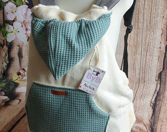 Puede incluir: Un portabebés blanco con una capucha y un bolsillo de tejido tipo gofre de color turquesa. El portabebés tiene un ribete de cuero marrón y una etiqueta que dice "ByB Bebés y Bolsos Atelier".