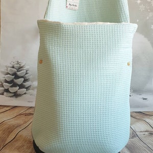 May include: A light blue waffle weave baby carrier cover with a white trim and two gold buttons. The cover has a tag that says "ByB Atelier".