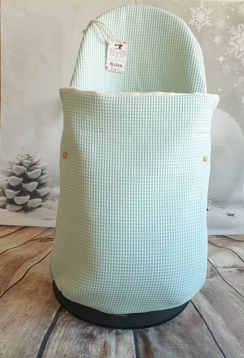 May include: A light blue waffle weave baby carrier cover with a white trim and two gold buttons. The cover has a tag that says "ByB Atelier".