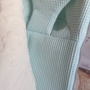 May include: A light blue waffle weave baby carrier with a white fleece lining. The carrier has a zipper closure and a brown leather tag.