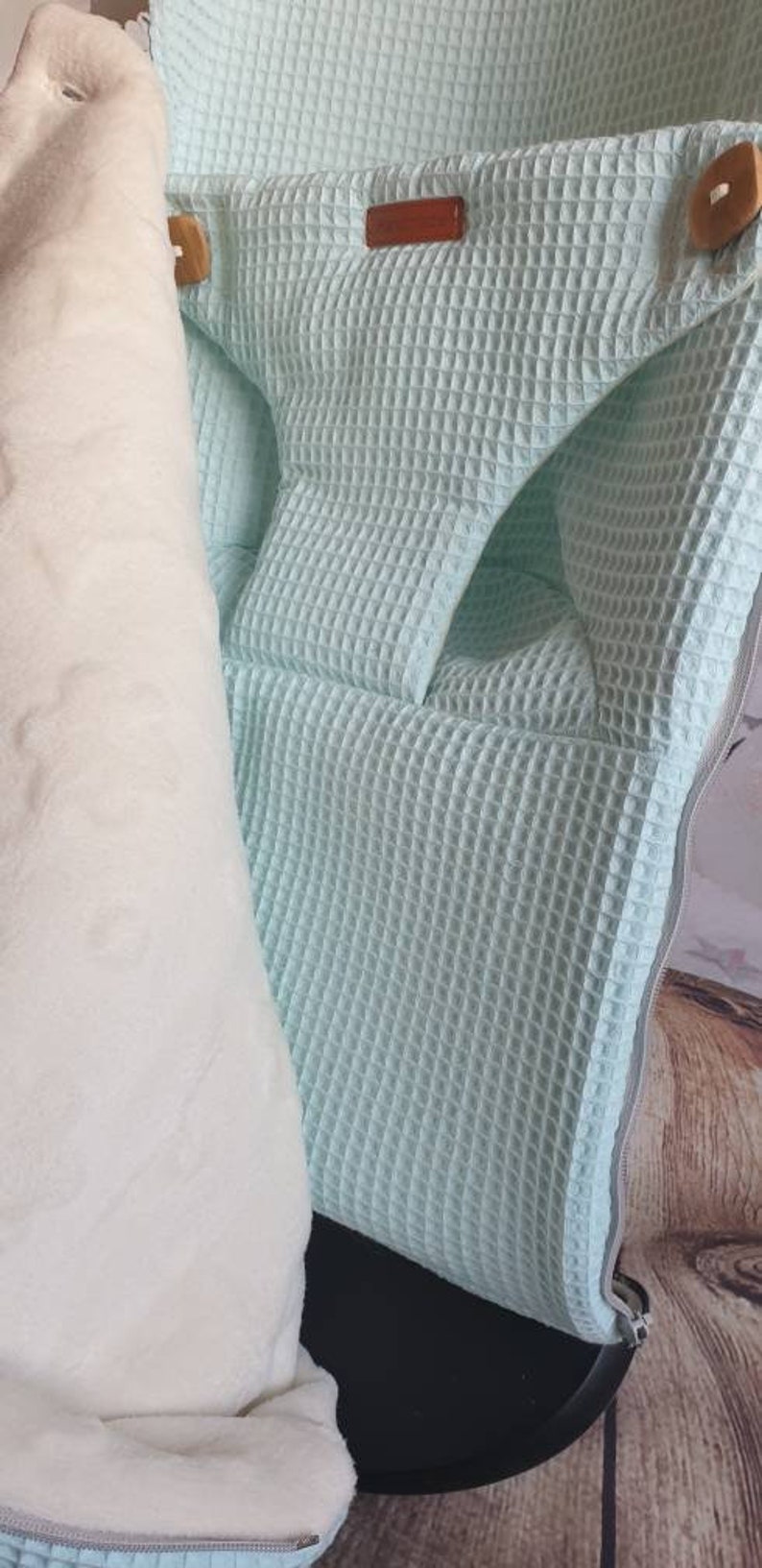 May include: A light blue waffle weave baby carrier with a white fleece lining. The carrier has a zipper closure and a brown leather tag.