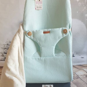 May include: A light blue waffle weave baby chair cover with a brown leather strap and wooden buttons. The cover is on a black base with the text "BabyBj&ouml;rn" printed on it. The cover has a tag attached with the text "ByB Atelier".