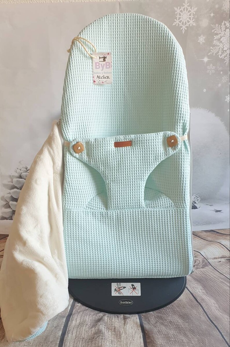 May include: A light blue waffle weave baby chair cover with a brown leather strap and wooden buttons. The cover is on a black base with the text "BabyBj&ouml;rn" printed on it. The cover has a tag attached with the text "ByB Atelier".