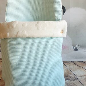 May include: A light blue waffle weave baby carrier cover with a white fleece lining and a wooden button. The cover has a tag that says "ByB Atelier".