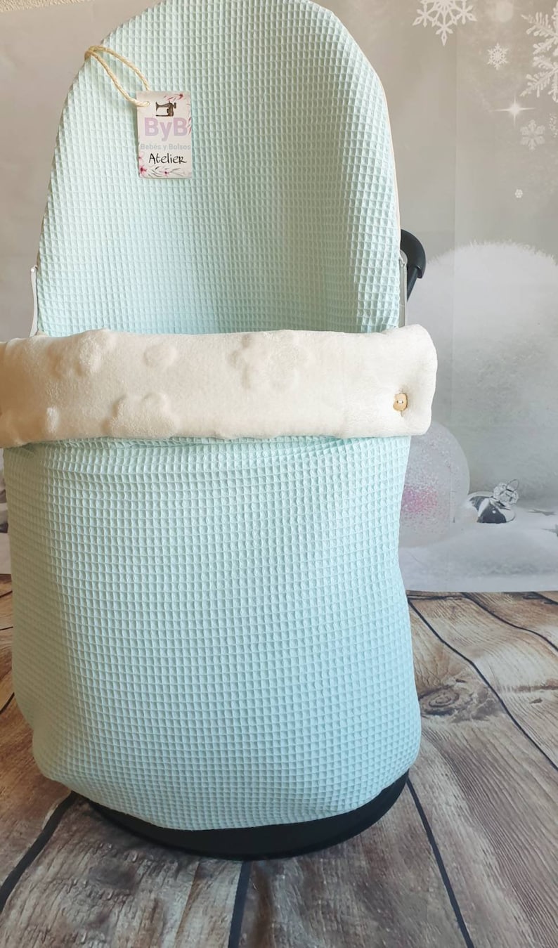 May include: A light blue waffle weave baby carrier cover with a white fleece lining and a wooden button. The cover has a tag that says "ByB Atelier".