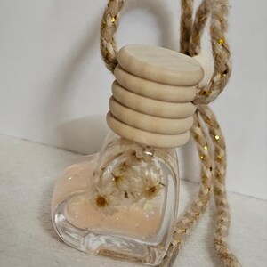 May include: A heart-shaped glass bottle with a pink liquid and white flowers inside. The bottle is attached to a beige rope with gold accents.