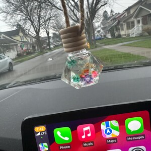 May include: A clear glass car air freshener with a wooden top and a string for hanging. The air freshener is filled with dried flowers and is hanging from the rearview mirror of a car. The car's infotainment system is visible in the background, showing the time, date, and icons for phone, music, maps, messages, calendar, and settings.