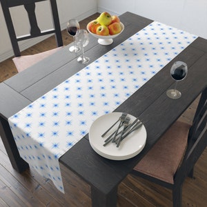 May include: White table runner with a blue starburst pattern. The runner is on a dark wood table with a bowl of apples, two wine glasses, and a stack of white plates with silverware.