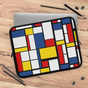 May include: Laptop sleeve featuring a geometric design with red, blue, yellow, and white rectangular shapes on a black background. The sleeve has a black zipper and is placed on top of a laptop on a wooden surface. Pencils and paper clips are also visible.