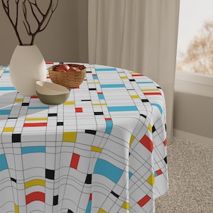 May include: A round tablecloth with a geometric design featuring a grid pattern with black lines and colorful blocks of red, yellow, blue, and black. The table is set with a white vase, a small bowl, and a basket of fruit.