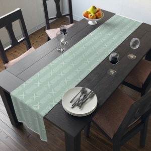 May include: A light green table runner with a white pineapple pattern. The runner is on a dark wood table with two chairs and a bowl of fruit.