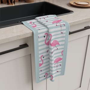 May include: A blue and white striped kitchen towel with a pink flamingo pattern. The towel is hanging on a white cabinet door.